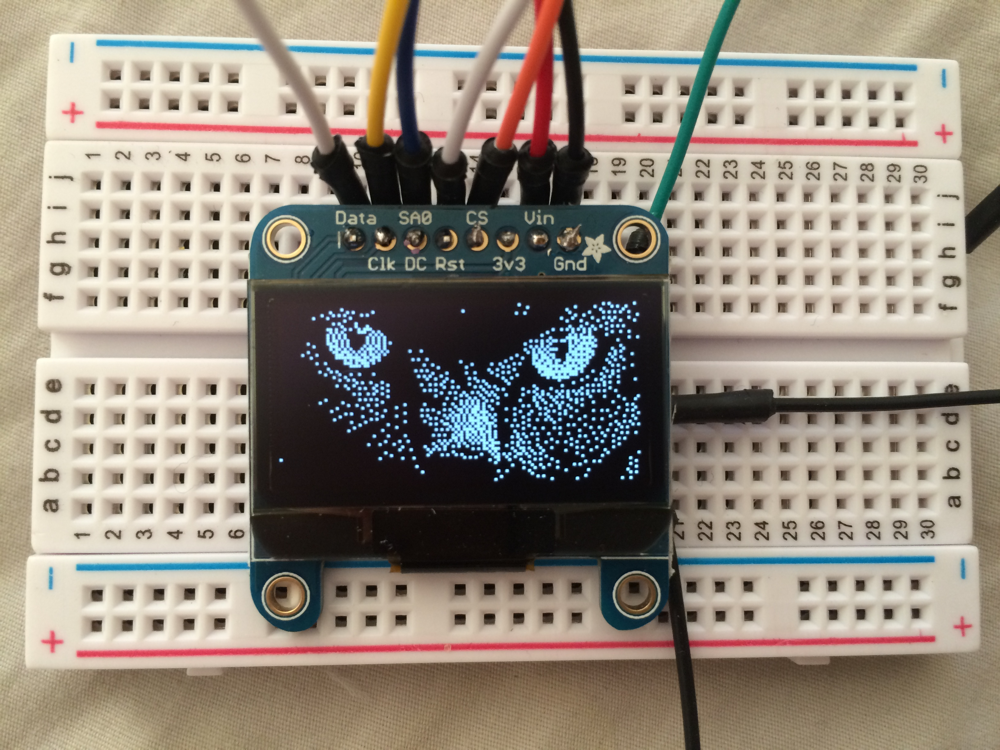 oled screen on a breadboard, displaing a pixelated monchrome picture of a cat's face